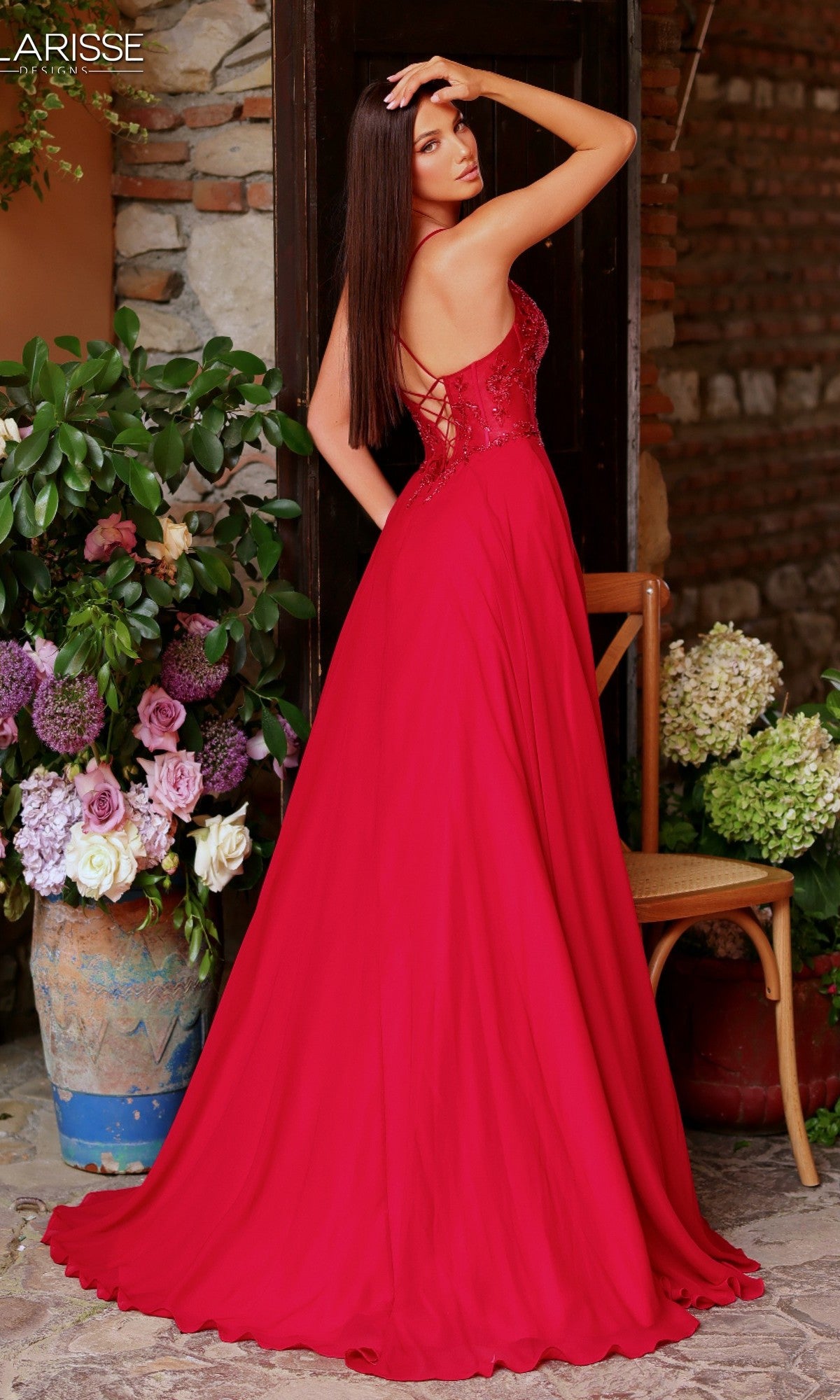 A woman wearing the Long Prom Dress: Clarisse Designs 62146, a red sleeveless gown with a chiffon A-line skirt, stands before a stone wall and large floral arrangement, glancing over her shoulder.