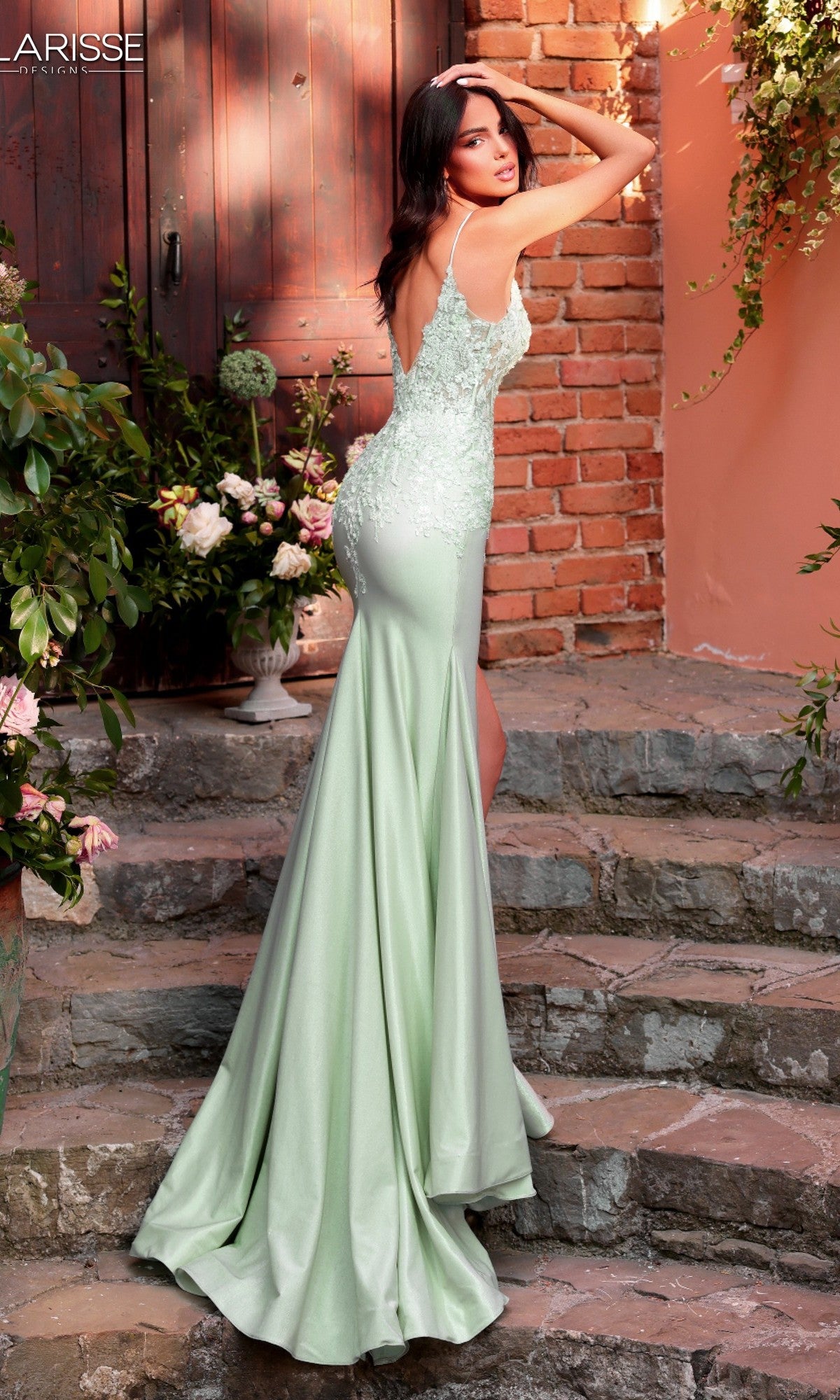 A woman poses on stone steps in front of a brick wall and wooden door, surrounded by greenery and flowers, wearing the Long Prom Dress: Clarisse Designs 62160—a light green, floor-length gown with a corset bodice.