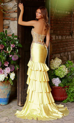A woman wearing the Long Prom Dress: Clarisse Designs 62162, featuring a strapless beaded corset bodice and ruffled yellow gown, stands in a doorway adorned with flowers and greenery against brick walls.