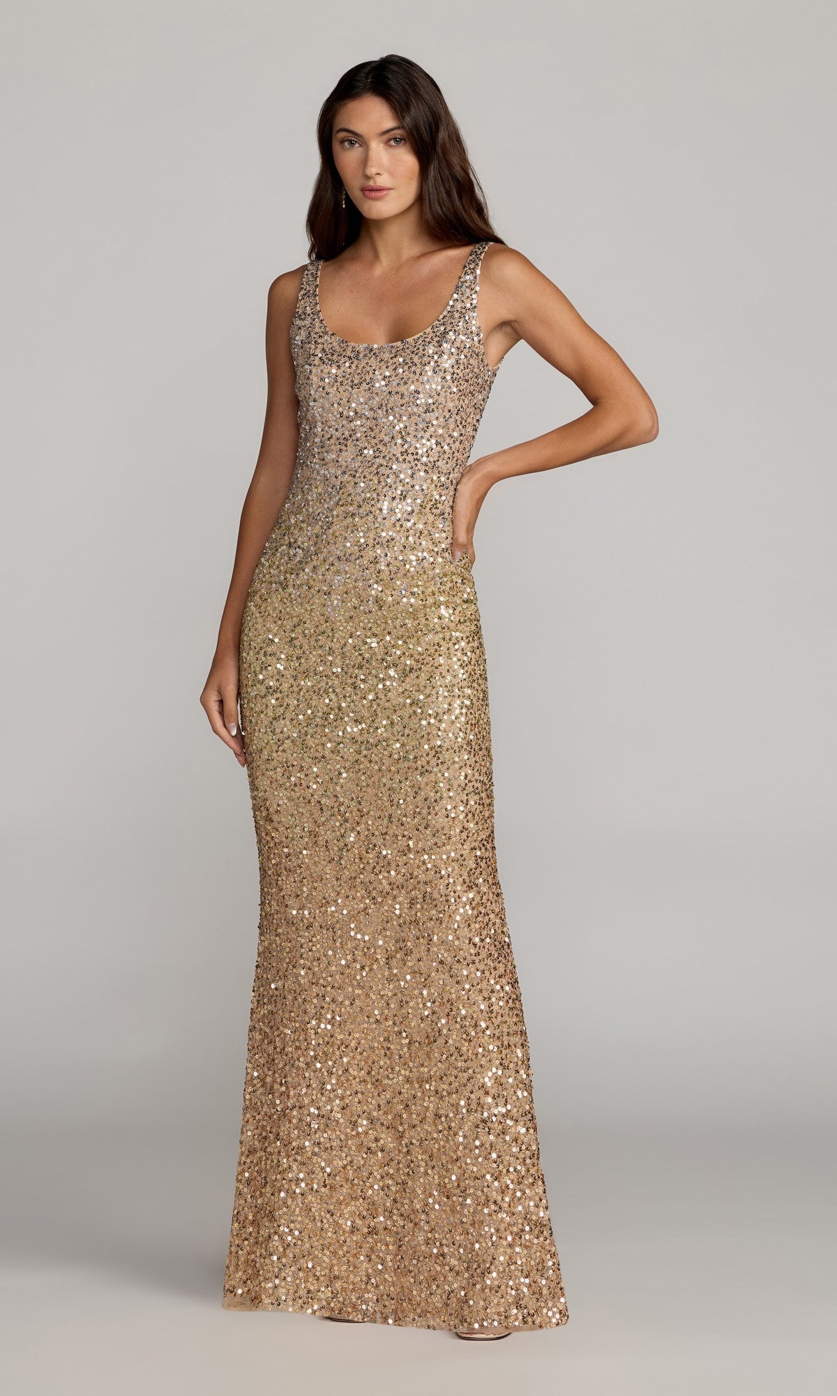 A woman stands with her hand on her hip against a plain light gray background, wearing the Mac Duggal 6219 Long Formal Dress in shimmering gold, featuring sleeveless styling and sequin embellishments that extend to the floor-length hem.