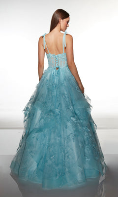 A woman stands with her back to the camera, wearing the Alyce 62199 long prom dress—a light blue gown with a corset bodice, floor-length tulle ballgown skirt, and delicate floral details—against a plain background.