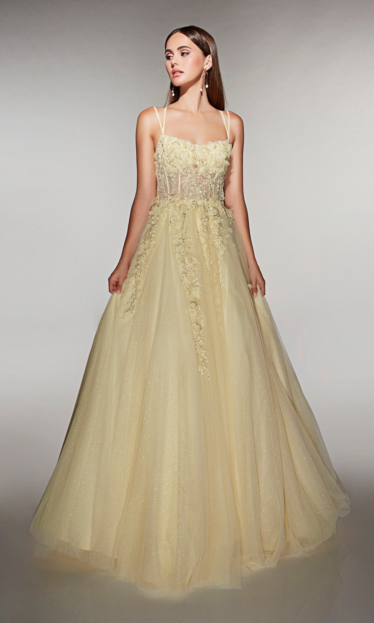 A woman models the Alyce 62203 long prom dress in light yellow, featuring floral appliqué, while standing against a plain gray background.