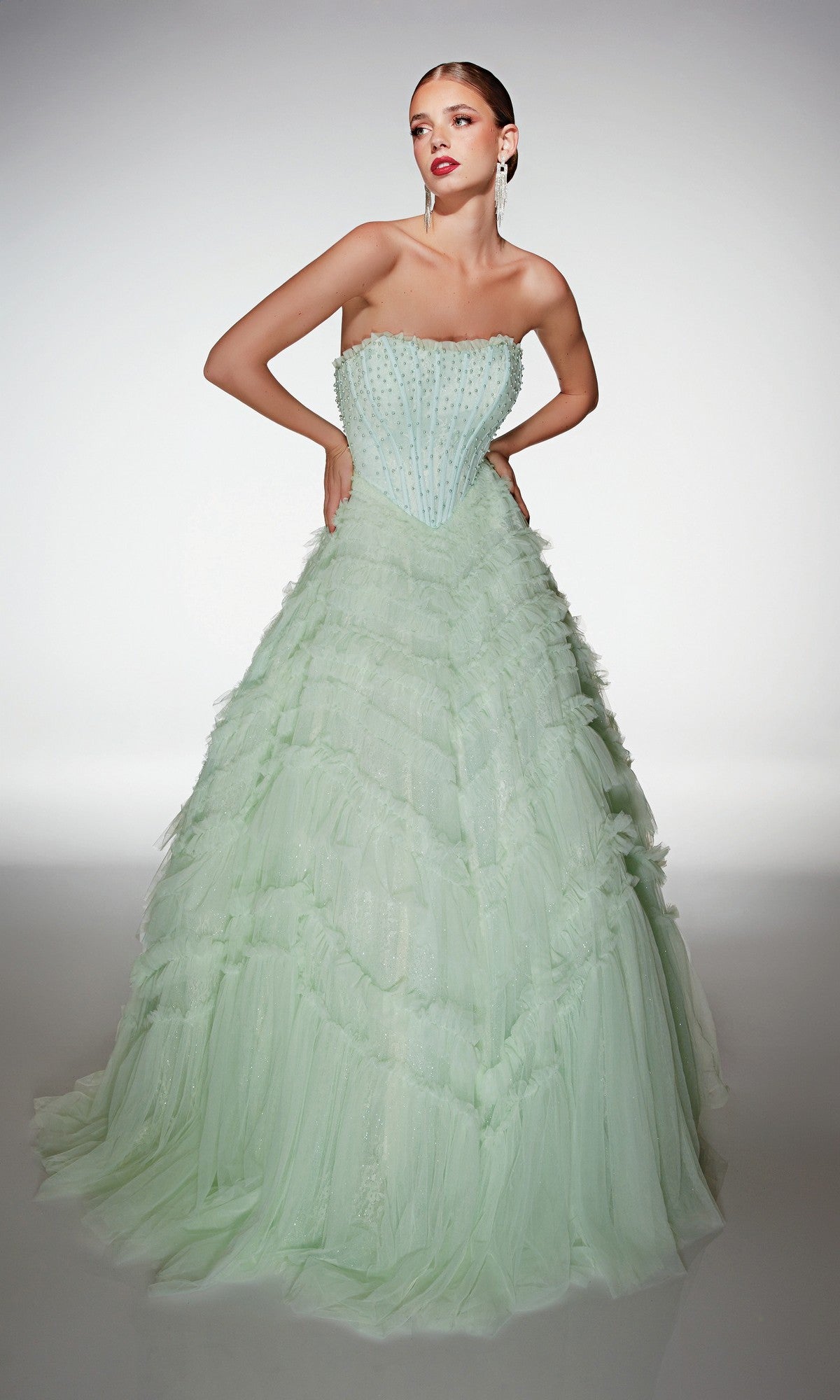 Wearing the Alyce 62208 long prom dress in mint, a woman stands against a plain backdrop. The strapless gown features beaded embellishments on the fitted bodice and a tiered tulle ruffle skirt.