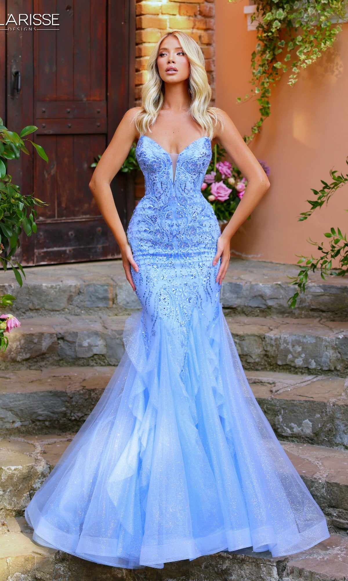 A woman in the sky blue Clarisse Designs 62258 long prom dress stands on stone steps outdoors, her glitter tulle skirt framed by a wooden door and flowers. The strapless mermaid gown features beaded embroidery.