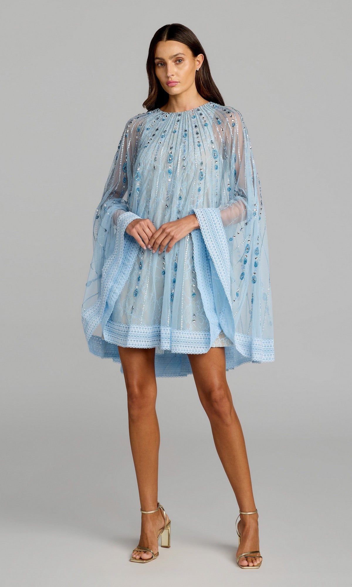 A woman stands against a plain gray background wearing the Mac Duggal 6226 short party dress in powder blue nude, featuring sheer cape sleeves with sequin embellishments and long, wide sleeves, paired with gold high-heeled sandals.