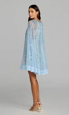 A woman stands in profile against a plain background, wearing the Mac Duggal 6226 short party dress—a sheer, light blue hand-beaded design with cape detail—paired with gold high-heeled sandals.