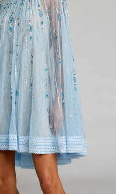A close-up of the Mac Duggal 6226 short party dress in blue lace, featuring beaded embellishments, a scalloped hem, and part of a person’s leg against a plain background.