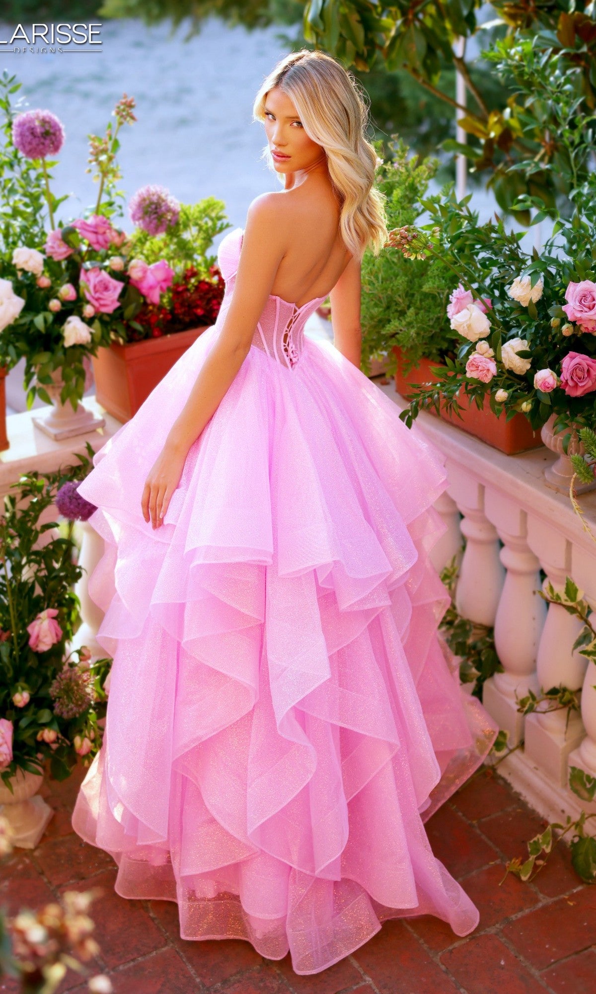 On a garden terrace surrounded by blooming flowers, a woman in the Clarisse Designs 62263 strapless glitter tulle prom ball gown stands gracefully, glancing over her shoulder.