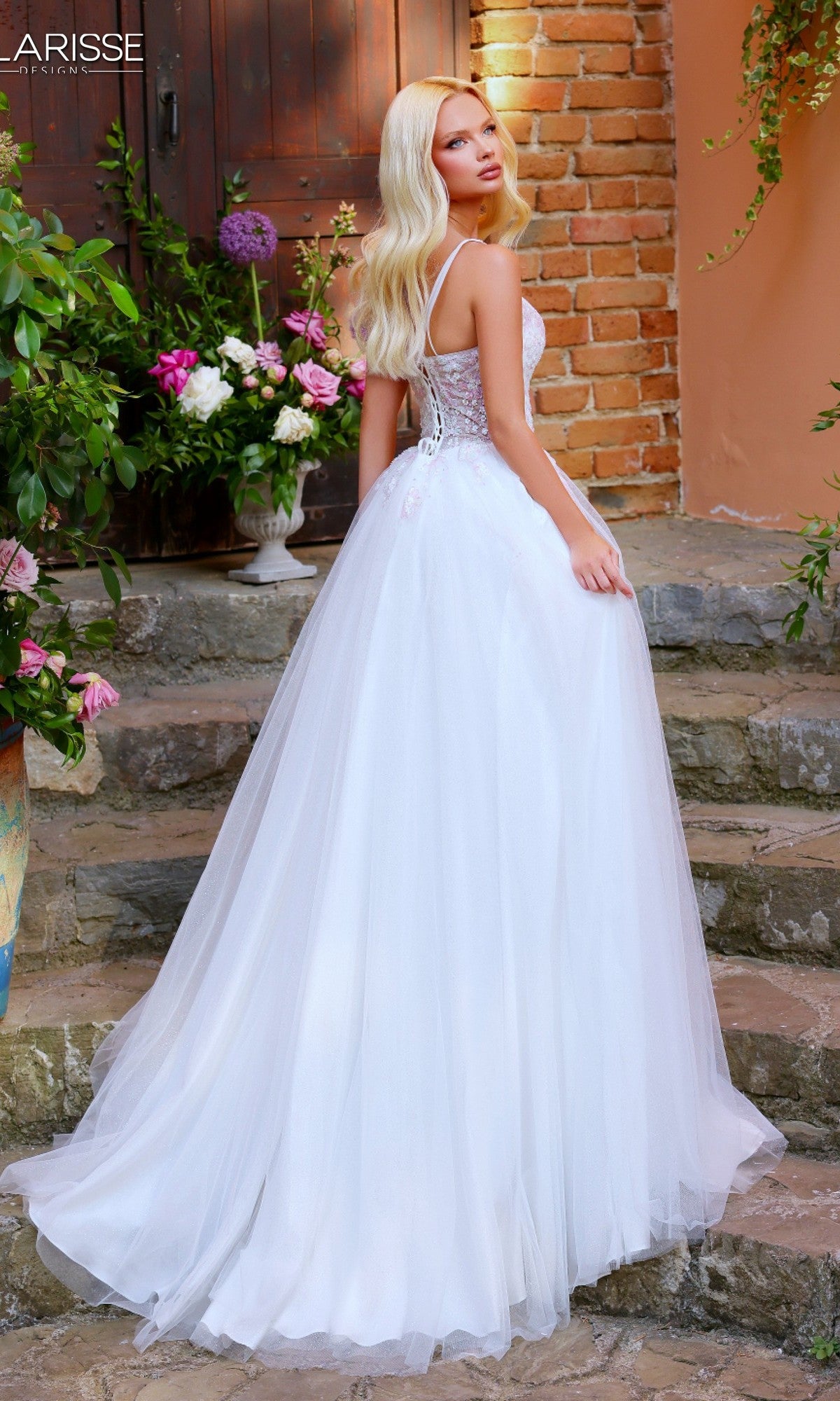 Wearing the Clarisse Designs 62314, a woman in a long white prom dress with a beaded embroidered bodice stands on stone steps outside, glancing over her shoulder amid flowers and a brick wall for an elegant look.