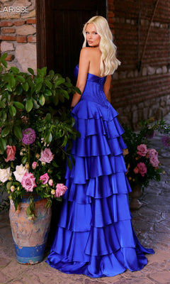 A woman with long blonde hair models the Clarisse Designs 62330 long prom dress, featuring a strapless satin corset bodice and a detachable ruffled royal blue tiered skirt, standing by a door with large potted flower arrangements.