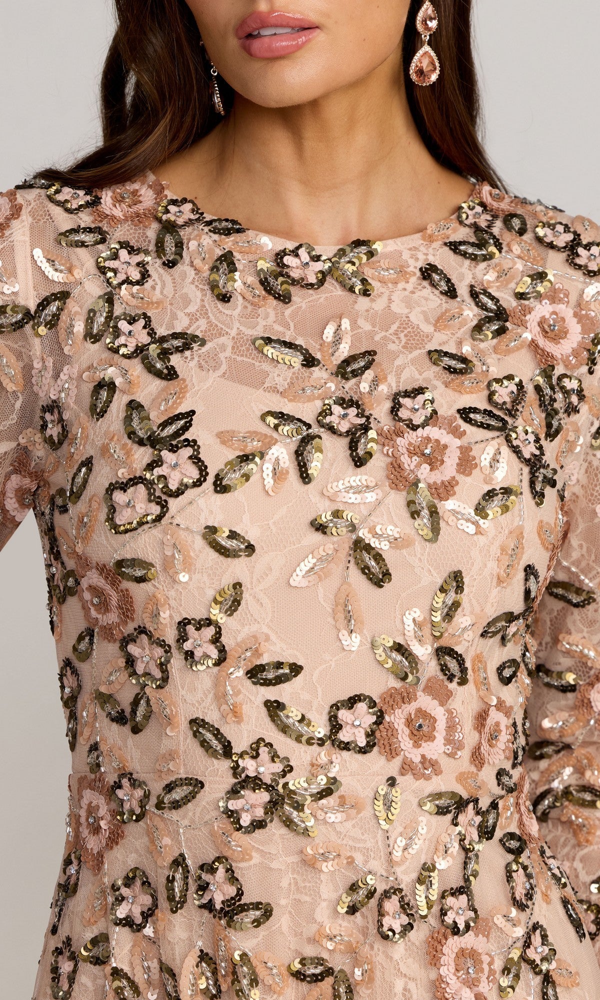A woman wearing the Mac Duggal 6239 Long Formal Dress, a sheer blush gown embellished with sequins and embroidered floral patterns in gold, pink, and black.