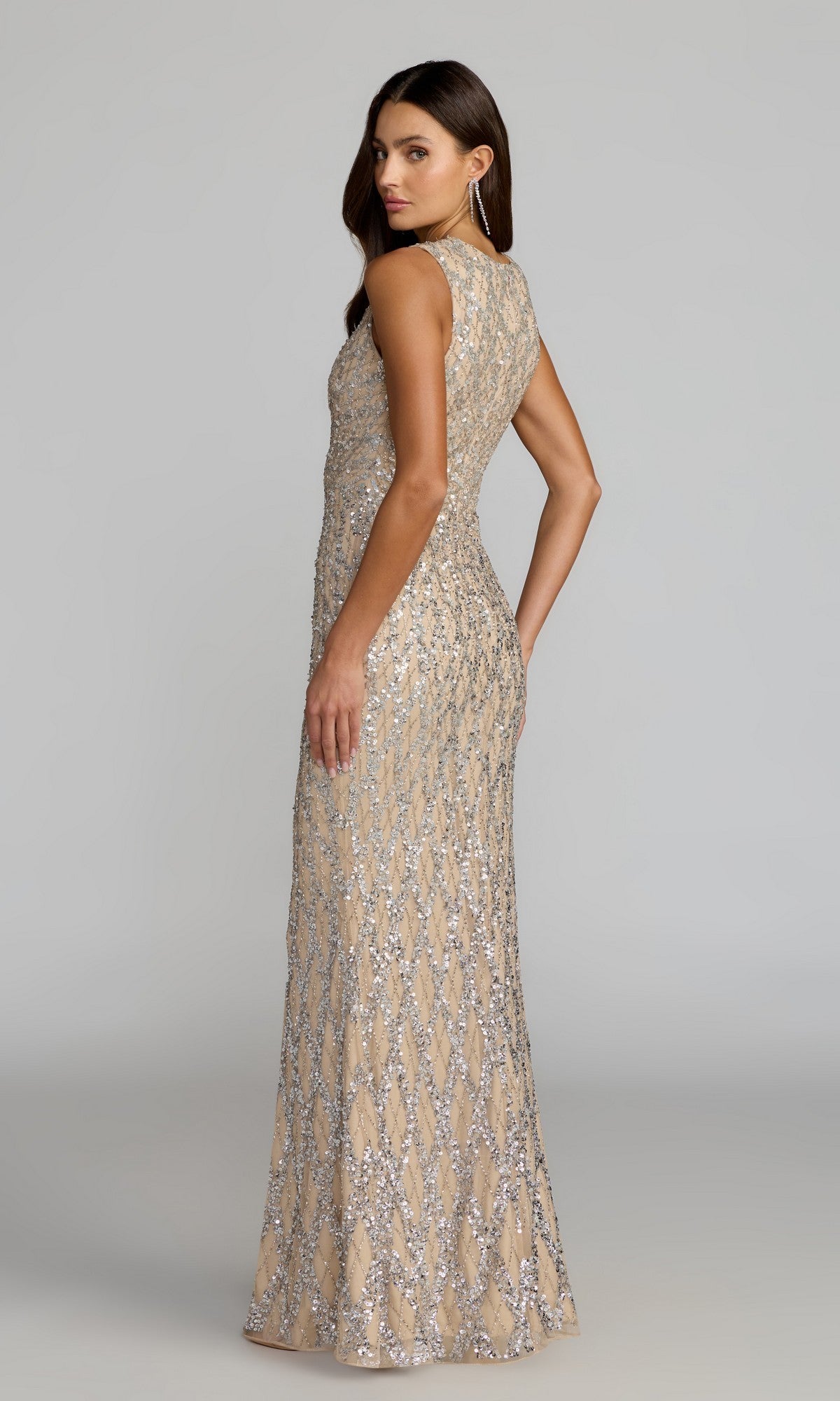 A woman stands facing backward with her head turned to the side, wearing the Mac Duggal 6249 long formal dress—a sleeveless, floor-length beige gown adorned with silver geometric sequin patterns.