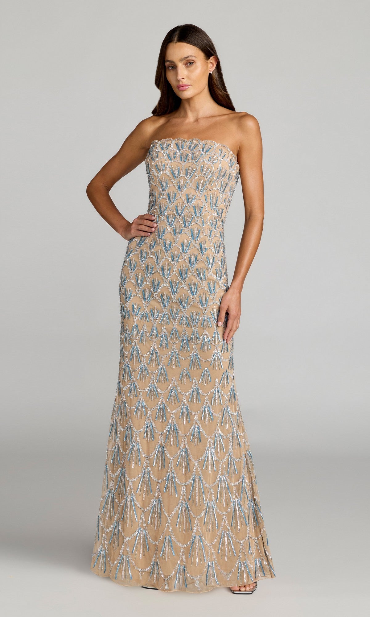 A woman poses elegantly with one hand on her hip against a plain background, wearing the Mac Duggal 6250 Long Formal Dress in Nude Silver—a strapless, floor-length evening gown intricately hand-beaded with shimmering silver and blue sequin patterns.