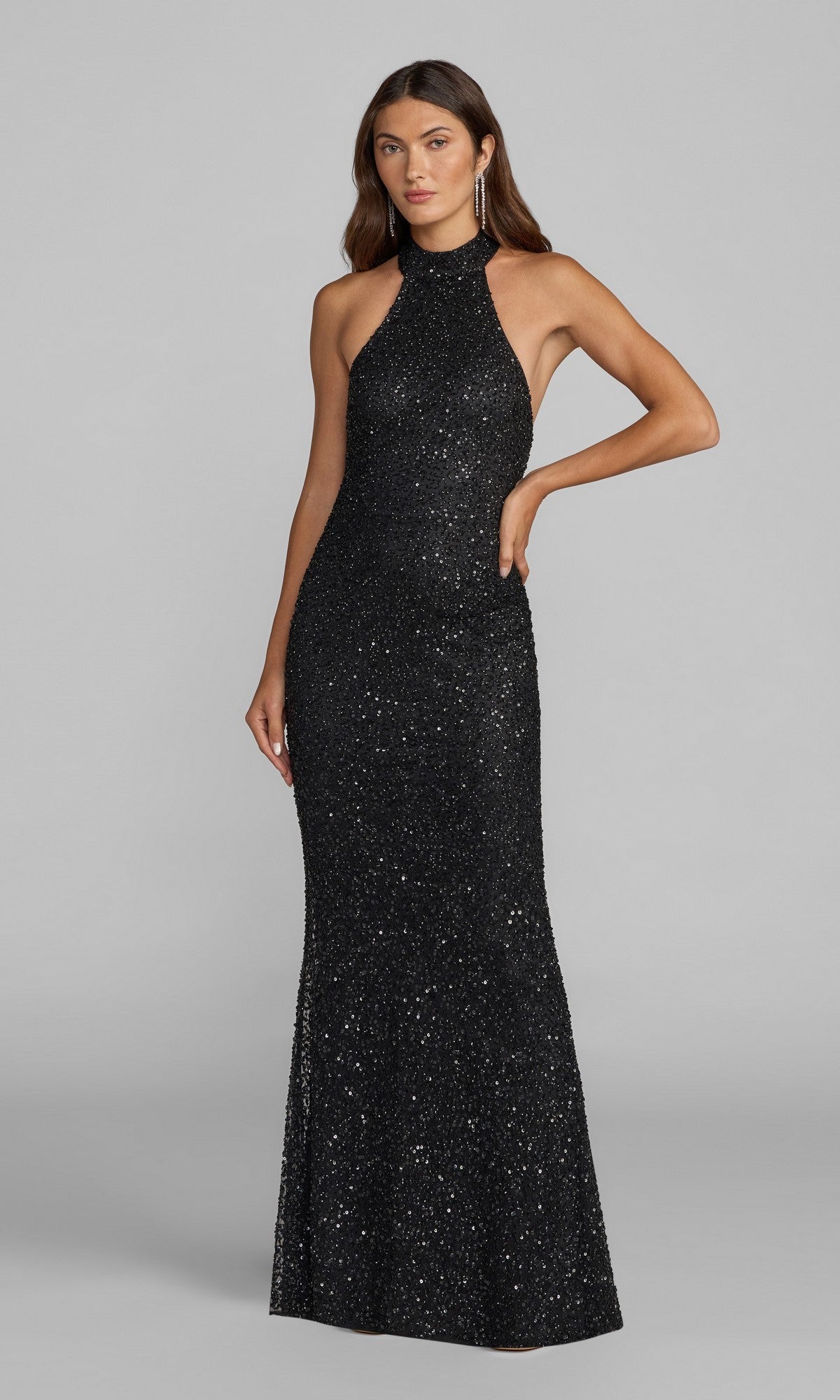 A woman stands with one hand on her hip against a plain gray background, wearing the Long Formal Dress: Mac Duggal 6259 in black, a sleeveless, halter-neck, floor-length sequin gown.