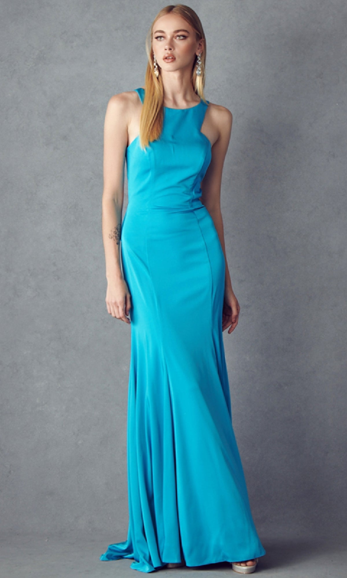 A woman poses against a gray backdrop wearing the Juliet 645, a sleeveless, floor-length turquoise prom dress with a racer front, paired with statement earrings.