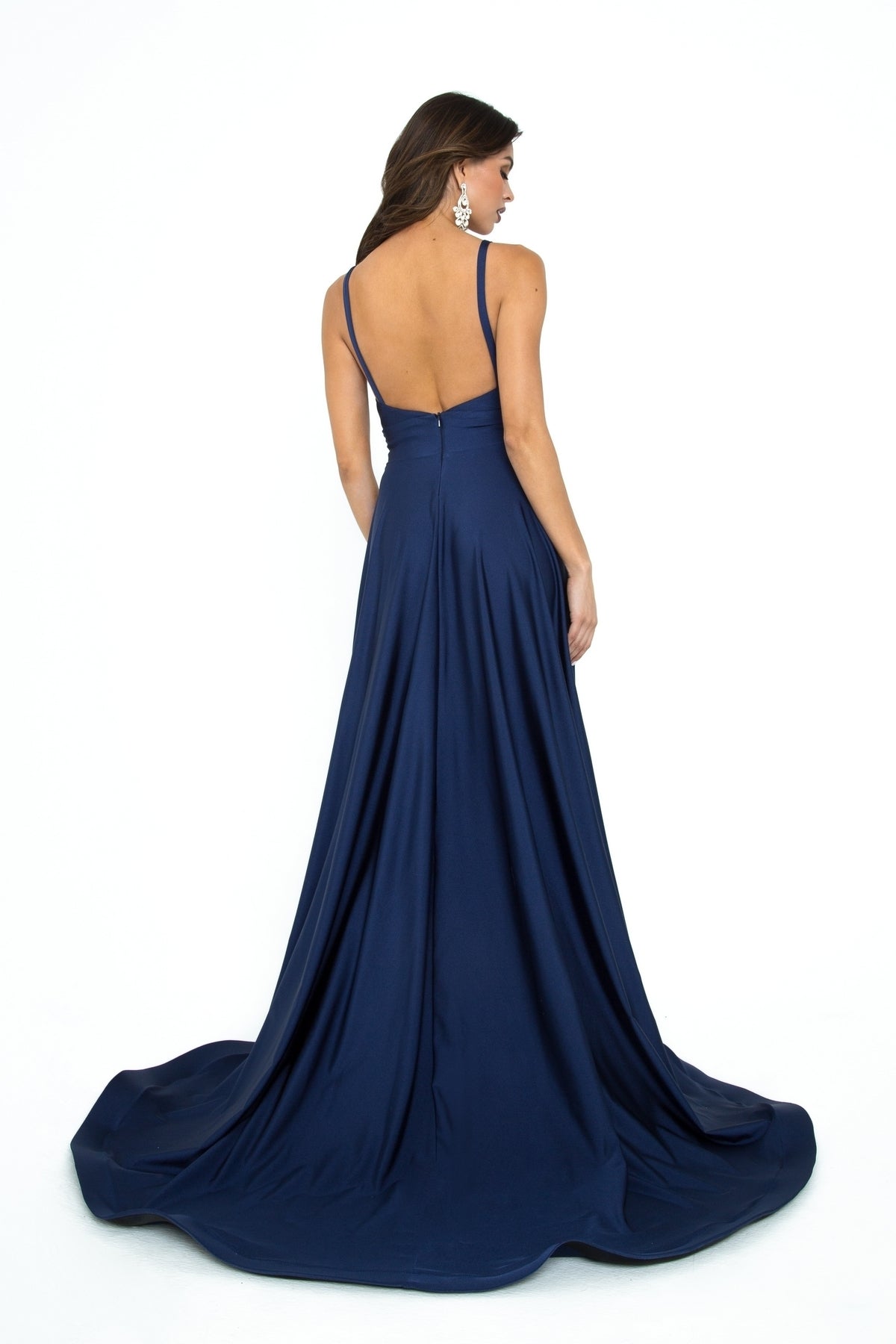 A woman stands with her back to the camera against a white background, wearing the Open-Back Long A-Line Prom Dress: Atria 6505H, a floor-length gown in deep blue with thin straps and a dramatic low back.