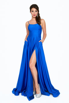 A woman stands facing forward, hands in the pockets of a bright blue Long A-Line Prom Dress with Pockets: Atria 6521H, featuring a thigh-high slit and paired with silver high-heeled shoes.