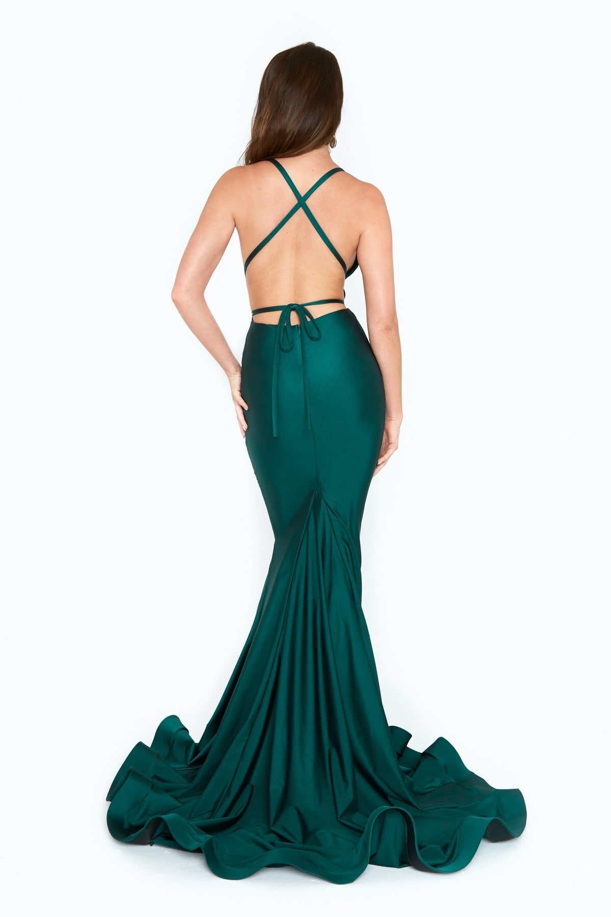 A woman stands against a white background wearing the emerald green Strappy-Back Long Mermaid Prom Dress (Atria 6530H) with crisscross straps and a flared train.