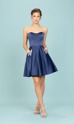 A person in the Eureka 6622 Strapless Short Wedding-Guest Dress—knee-length, dark blue with floral embroidered pockets—and silver open-toe heels stands against a light blue background.
