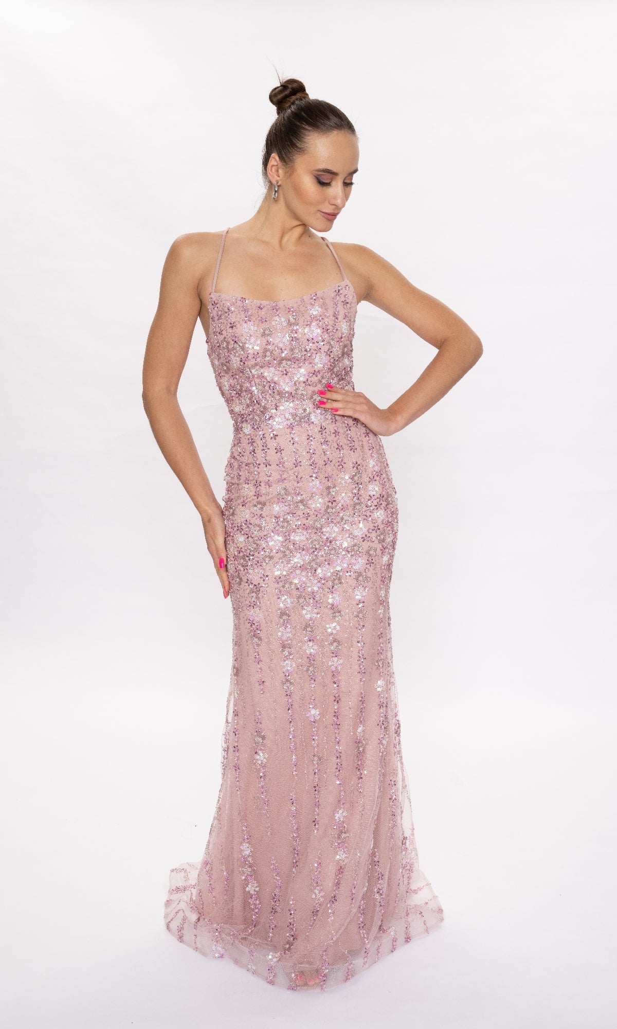 A woman wearing the Nina Canacci 6625 long formal dress in mauve pink with sequin and floral details stands against a white background, looking down with one hand on her hip.