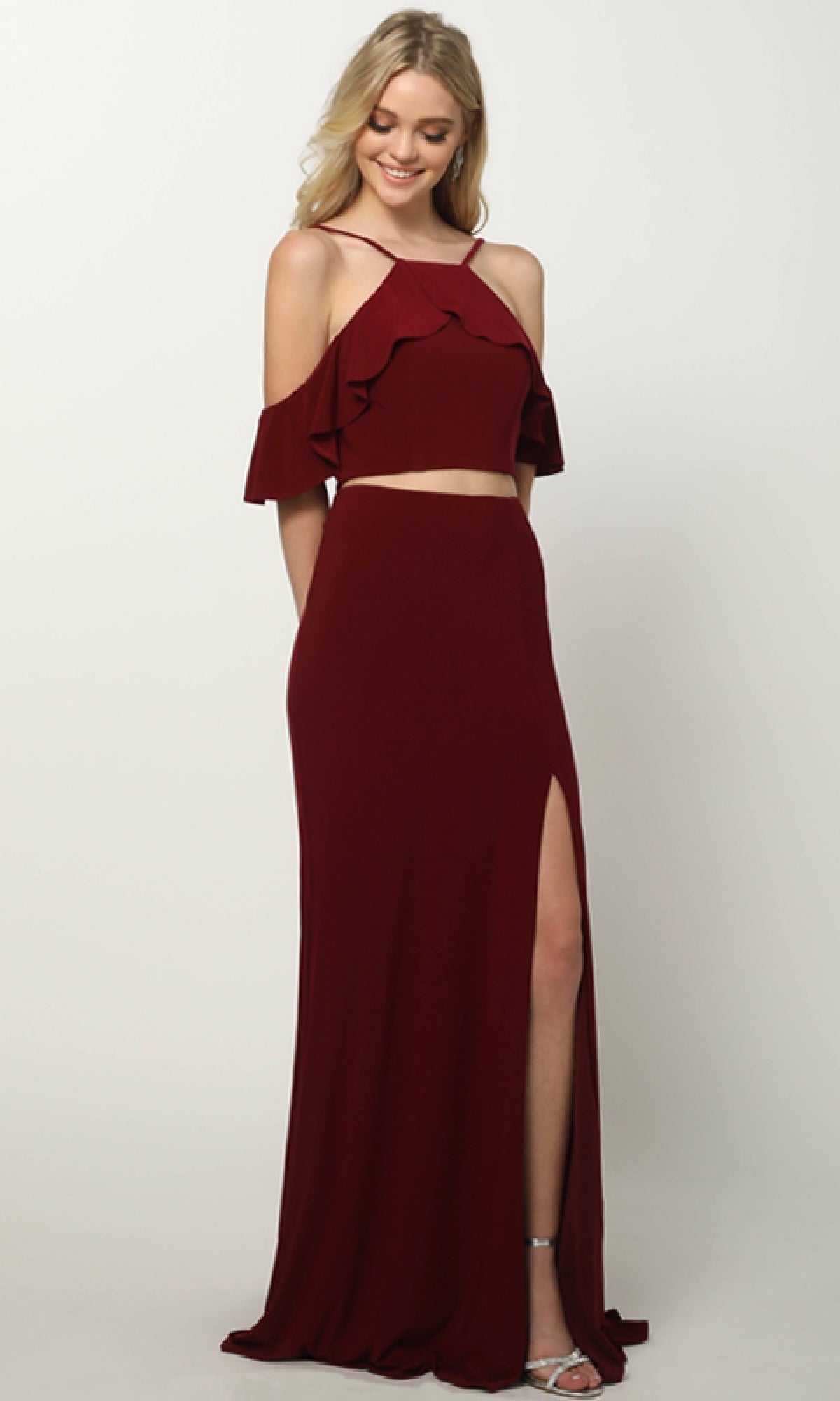 A woman models the Juliet 662 long prom dress, a burgundy two-piece with ruffled off-shoulder sleeves and a floor-length skirt featuring a dramatic leg slit, posed against a plain background.