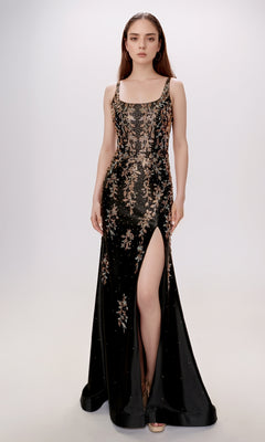 A woman stands against a plain light background wearing the Nina Canacci 6633 long formal dress—a sleeveless black gown with a square neckline, gold floral embroidery, and a high thigh slit.