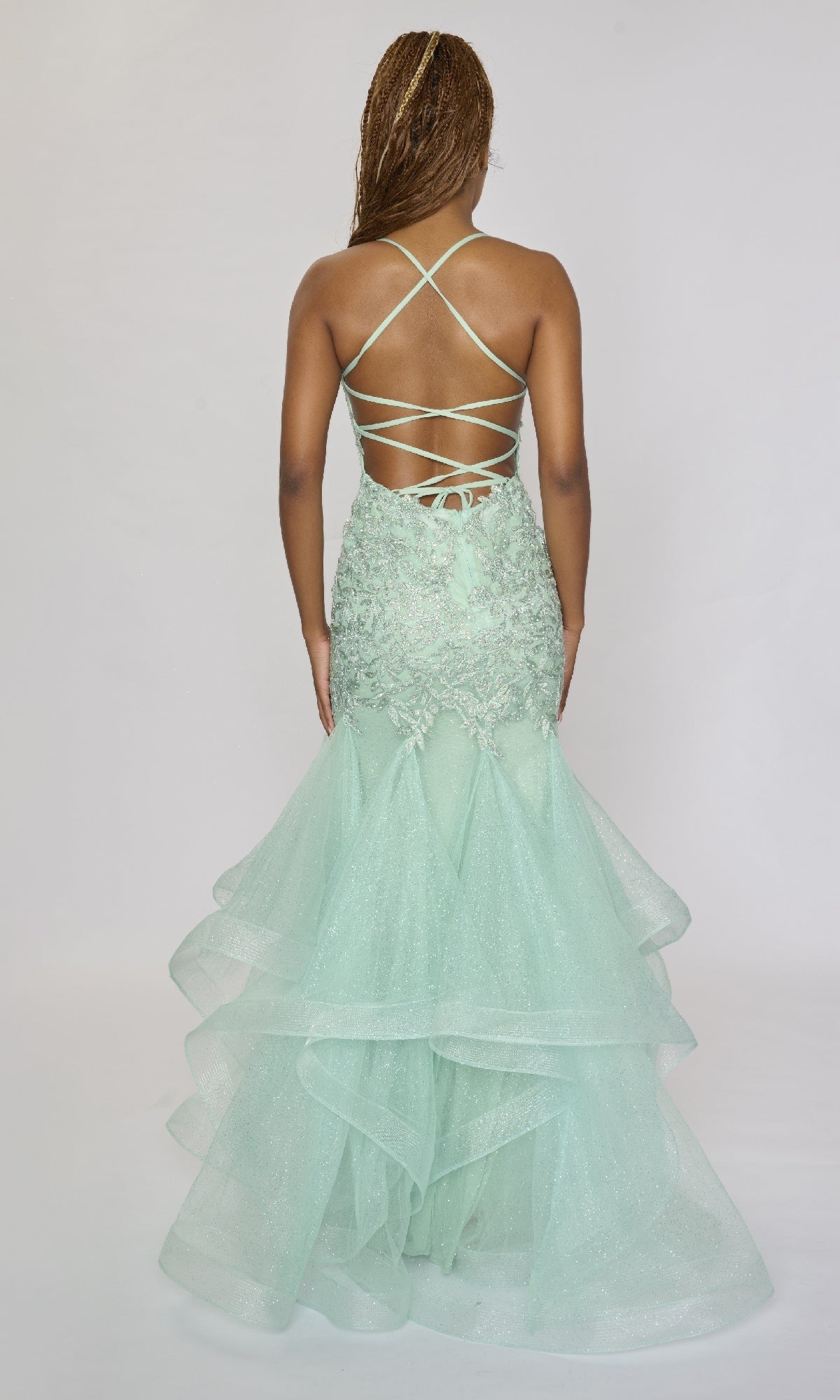 A woman stands facing away, wearing the Nina Canacci 6637 long formal dress in light green, featuring a mermaid silhouette, lace-up open back with crisscross straps, and a layered tulle skirt.
