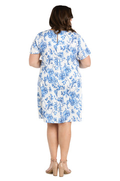 Woman standing with her back to the camera, wearing the R&M Richards 6674W Plus-Size Short Print Party Dress, featuring a white floral print with blue details and nude block-heeled shoes, against a plain white background.
