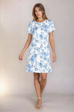 A woman wears the R&M Richards 6674 short sleeve party dress in blue and white floral and beige ankle-strap heels, standing against a plain background.