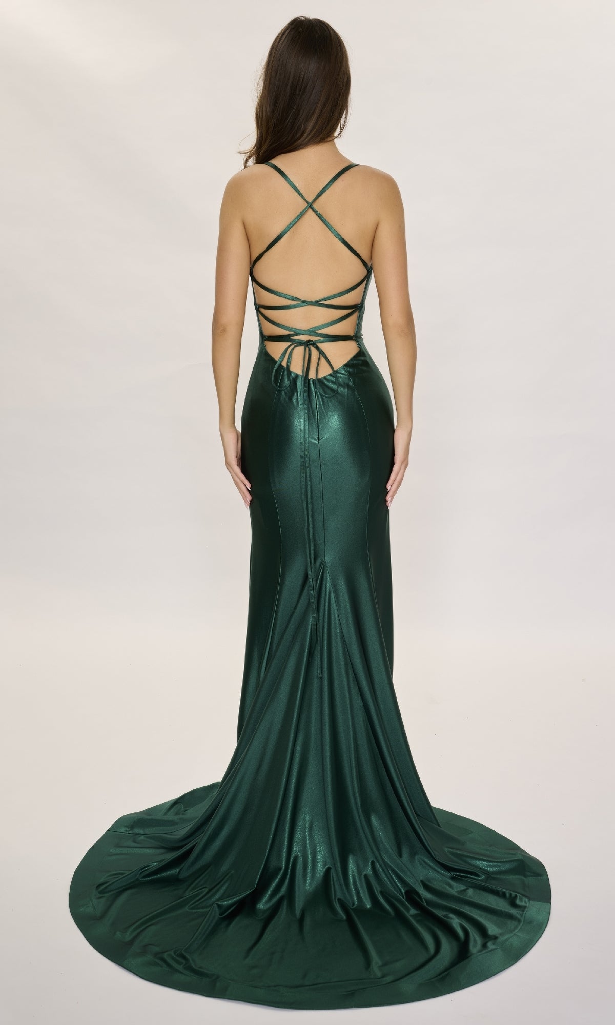 A woman stands facing away against a plain light background, wearing the Nina Canacci 6675— a dark green, floor-length formal dress with crisscross straps and a backless design.