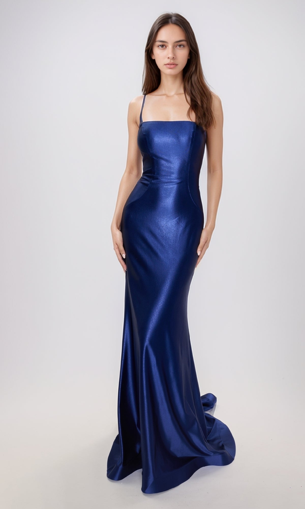 A woman stands against a plain background wearing the Nina Canacci 6675, a fitted, floor-length metallic formal dress with thin straps and a slight train.