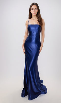 A woman stands against a plain background wearing the Nina Canacci 6675, a fitted, floor-length metallic formal dress with thin straps and a slight train.