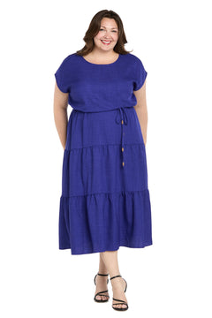 A woman smiles wearing the R&M Richards 6680W Plus-Size Indigo Casual Dress with short sleeves, waist tie, and tiered skirt, paired with black strappy sandals.