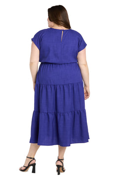 A woman models the Plus-Size Indigo Casual Dress: R&M Richards 6680W, featuring short sleeves and ankle-length tiered design, styled with black heeled sandals and shown from the back against a white background.
