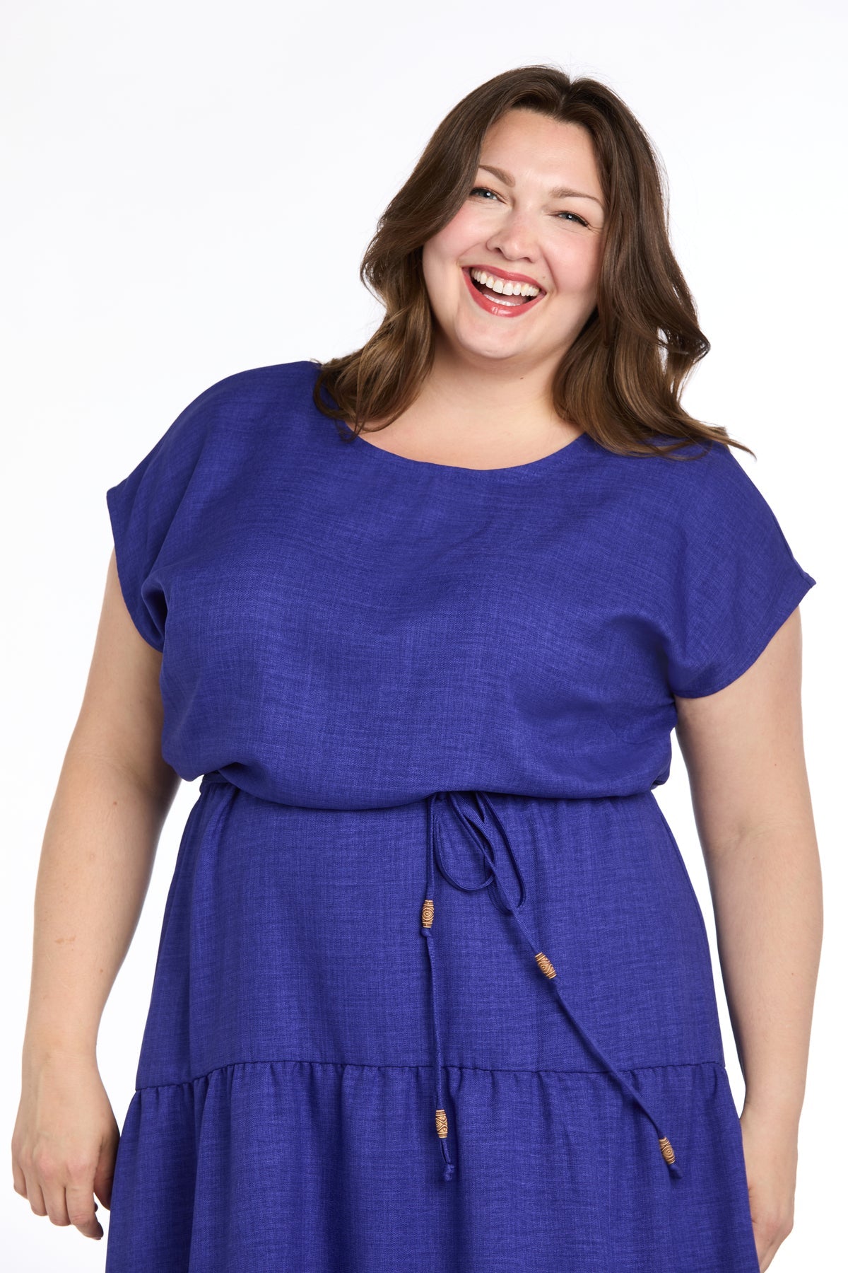 A woman with long brown hair smiles in the Plus-Size Indigo Casual Dress: R&M Richards 6680W, a short sleeve, belted knee-length blue dress, against a white background.