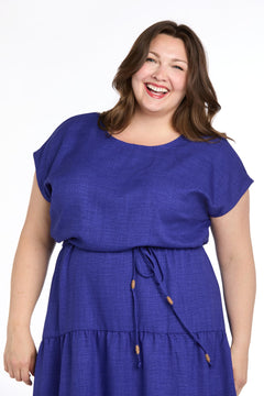 A woman with long brown hair smiles in the Plus-Size Indigo Casual Dress: R&M Richards 6680W, a short sleeve, belted knee-length blue dress, against a white background.