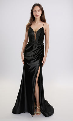 A woman stands against a plain light background wearing the Nina Canacci 6687—a black long formal dress with a plunging neckline, thin straps, thigh-high slit—and gold strappy heels.