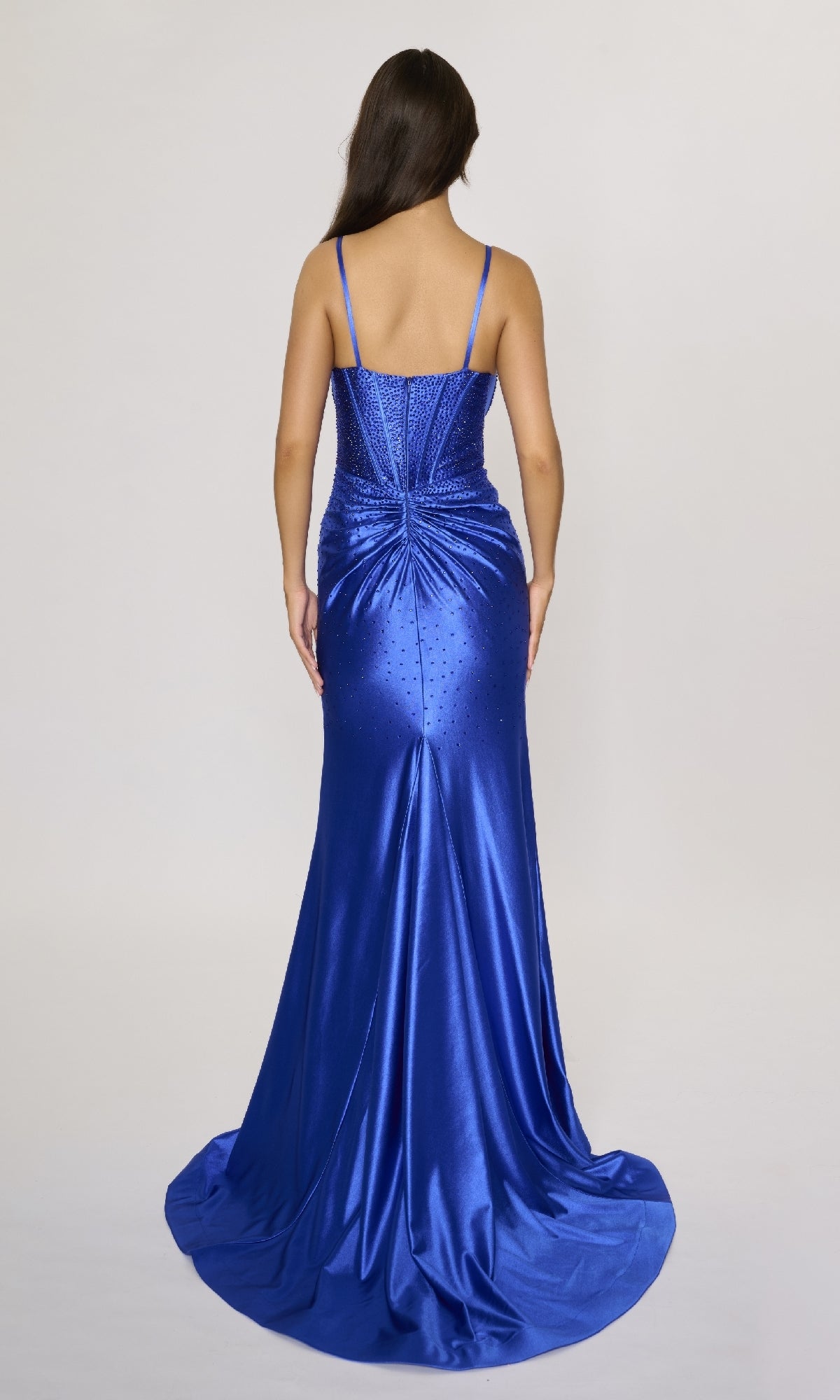A woman with long brown hair is photographed from behind wearing the Nina Canacci 6687 long formal dress in royal blue, featuring thin straps and rhinestone embellishments on the gathered back.