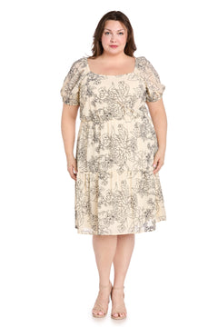 A woman faces forward in the Plus-Size Ivory Party Dress: R&M Richards 6688W, a knee-length ivory dress with black floral print and nude heels, shown against a plain white background—ideal as a floral print cocktail dress.