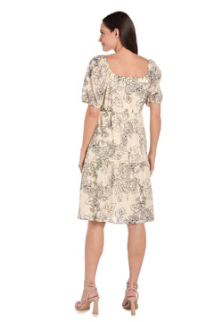 A woman faces away from the camera wearing the R&M Richards 6688P petite knee-length party dress, featuring a light-colored floral print, short puff sleeves, and a square neckline.