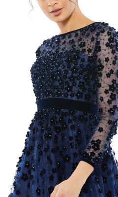 Woman in the Mac Duggal 67007 knee-length wedding-guest dress featuring navy blue floral embellishments, sheer sleeves, and appliqué details, shown from shoulders to mid-thigh against a white background.