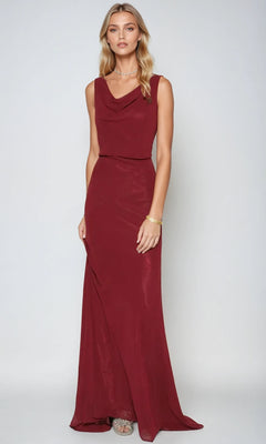 A woman stands against a plain backdrop wearing the Juliet 670, a long burgundy prom dress with a graceful cowl neckline and subtle jewelry.