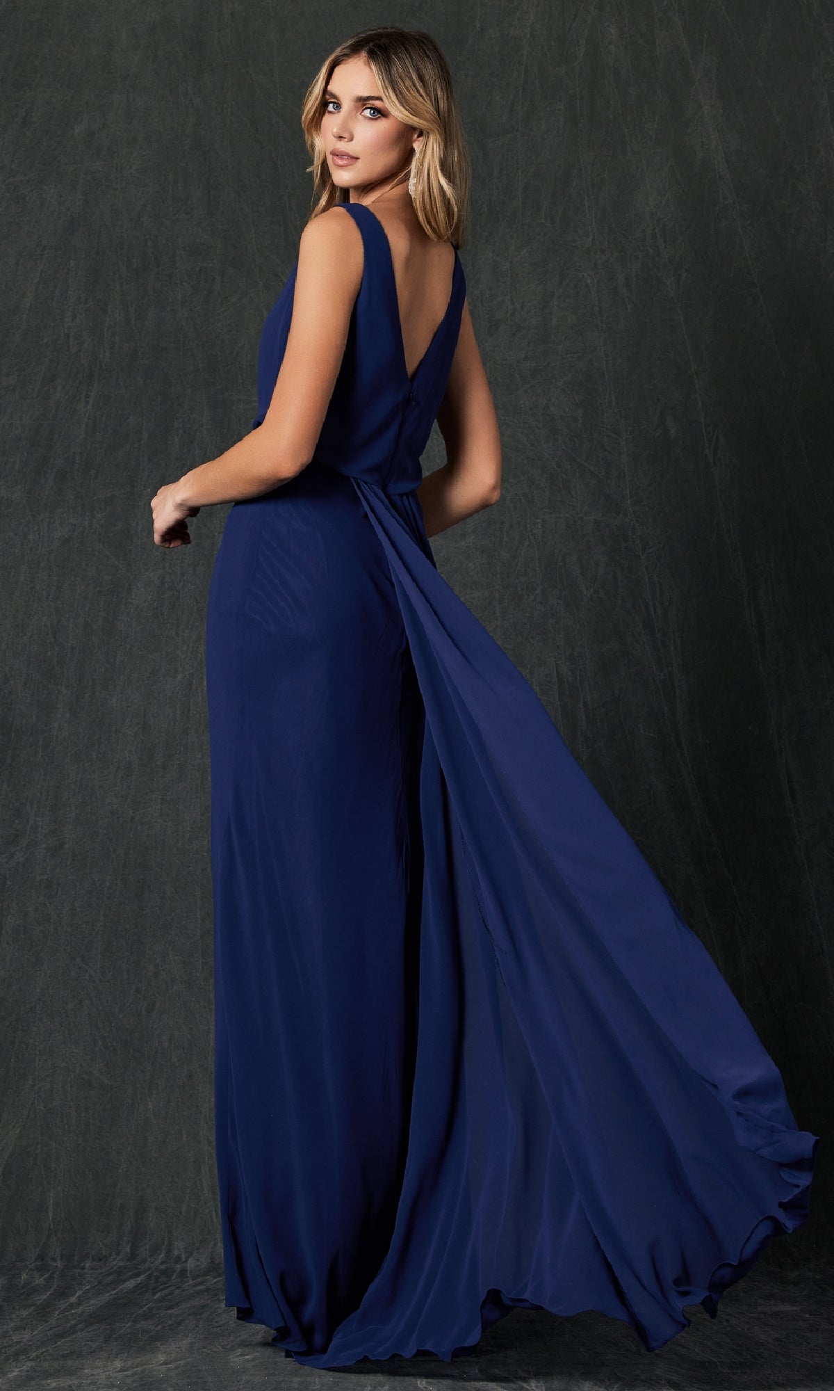 A woman models the Juliet 670 Long Prom Dress, featuring a sleeveless deep blue blouson bodice, V-back, and flowing train, set against a dark textured backdrop.