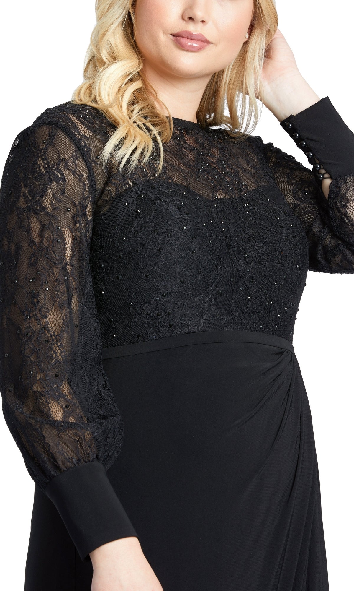 A woman poses with one hand in her hair, wearing the Mac Duggal 67143 plus-size black formal dress featuring sheer lace sleeves and beaded bodice detailing.