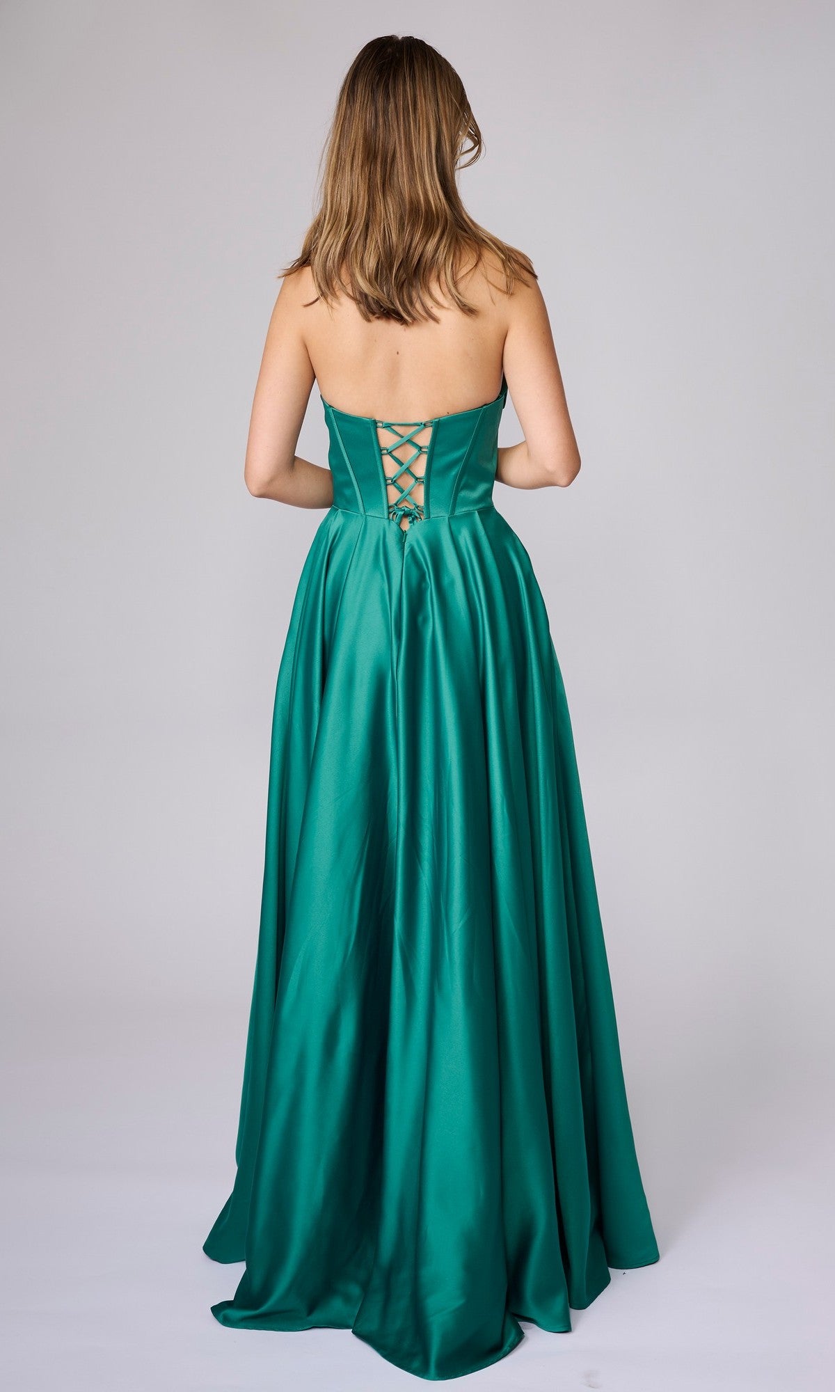 A woman stands facing away in the Nina Canacci 6718 long formal dress, a strapless emerald green satin A-line gown with a lace-up back and floor-length silhouette.