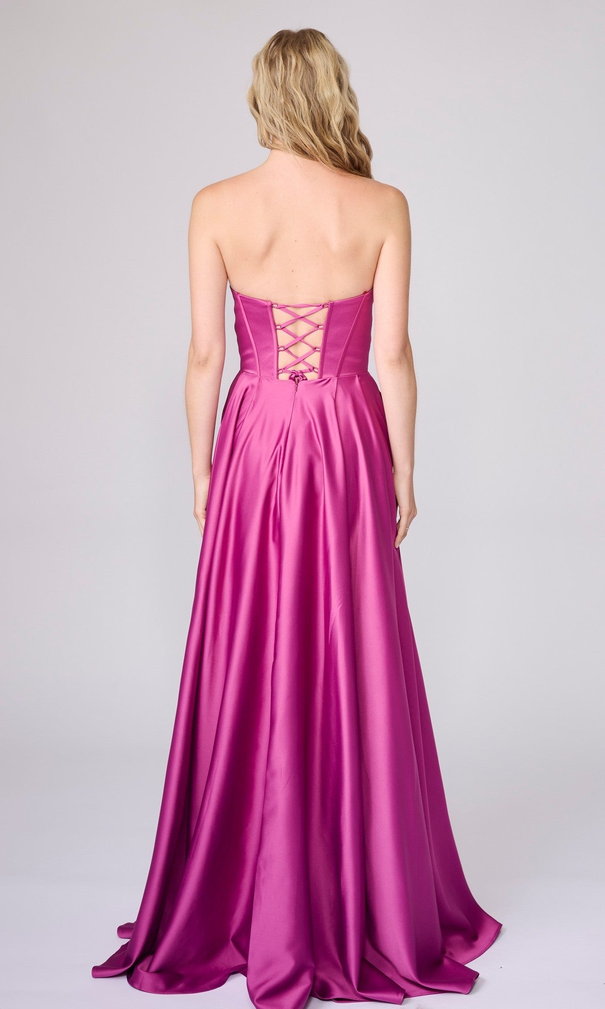 A woman with long blonde hair models the Nina Canacci 6718, a strapless magenta satin floor-length gown with a lace-up back, shown from behind against a plain light background.