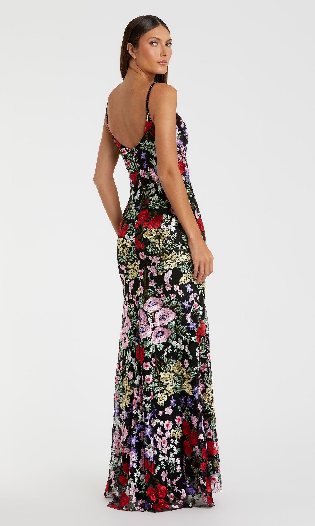 A woman stands against a plain white background, looking over her shoulder in the Mac Duggal 67473 Long Floral Formal Dress—a sleeveless, floor-length evening gown with an open back.