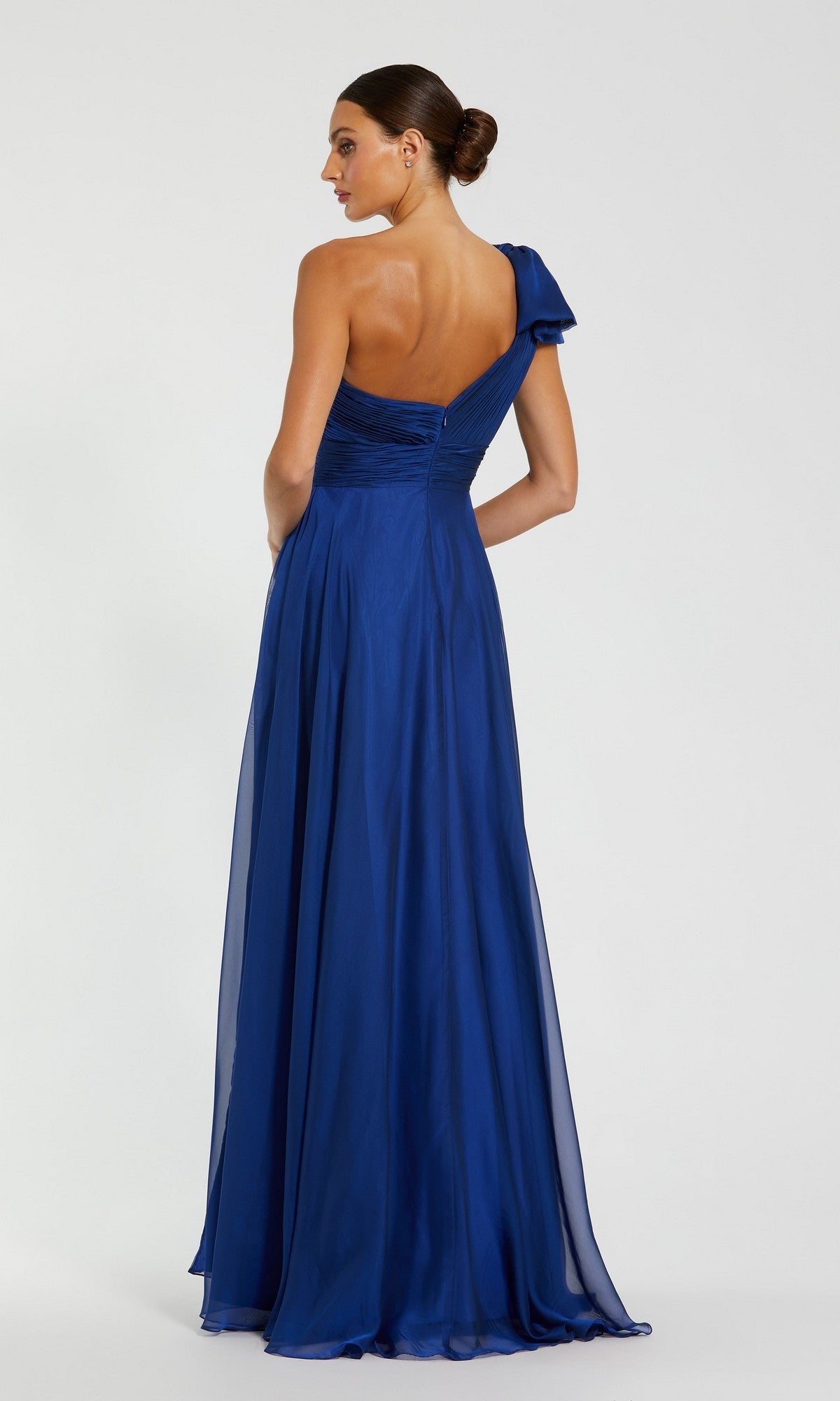 A woman stands with her back to the camera, wearing the Mac Duggal 67476 long one-shoulder prom dress in royal blue, against a plain light background.