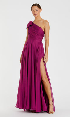 A woman models the Mac Duggal 67476, a magenta long one-shoulder prom dress with a high slit, standing against a white background to showcase the gown's elegant design.