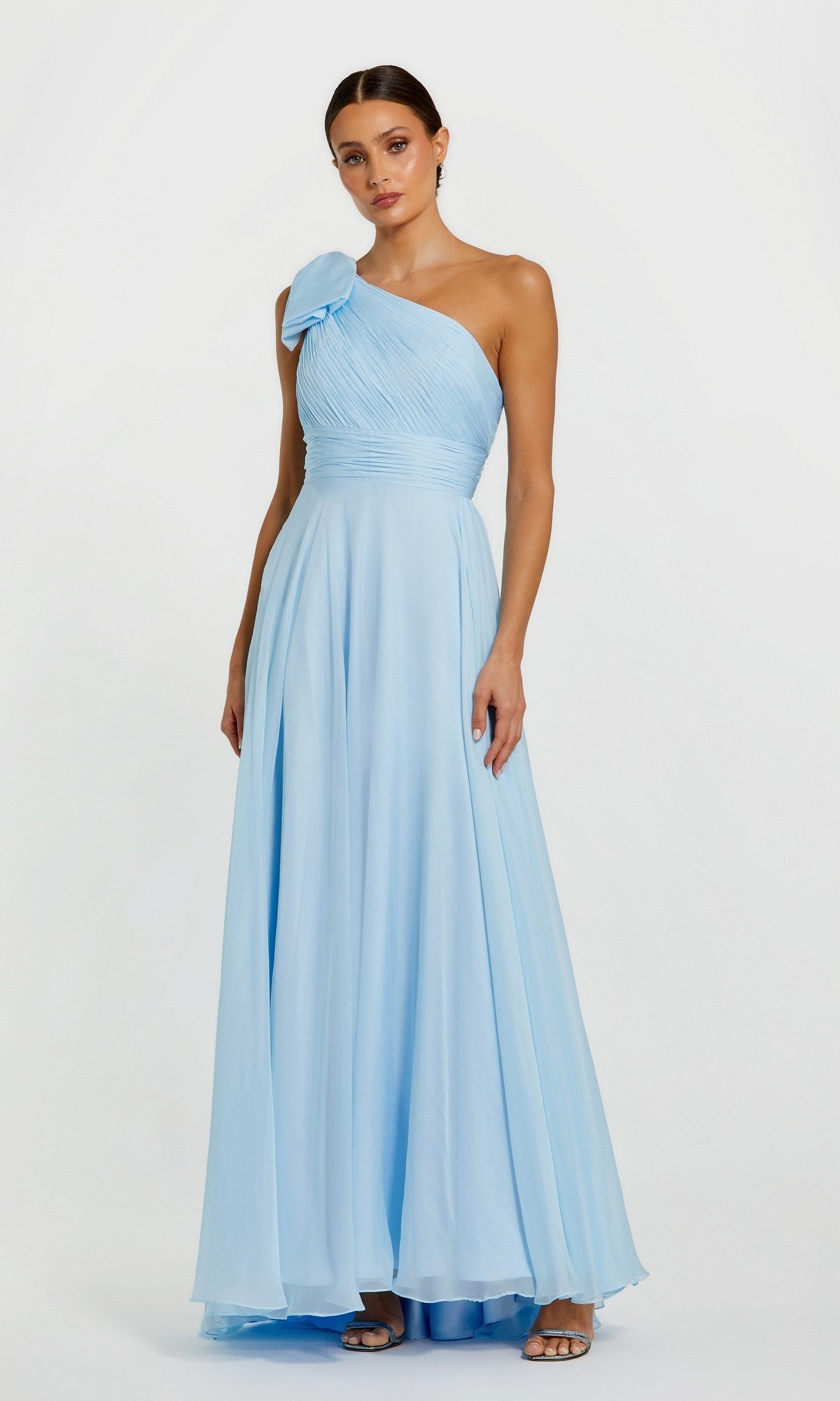 A woman models the Mac Duggal 67476 long one-shoulder prom dress in light blue with a bow detail, standing against a plain white background.